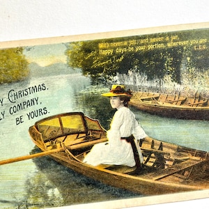 May include: A vintage postcard featuring a person in a white dress and yellow hat rowing a wooden boat on a river. The text reads: A MERRY CHRISTMAS, IN JOLLY COMPANY, BE YOURS. With never a jolt, and never a jar, Happy days be your portion, wherever you are.
