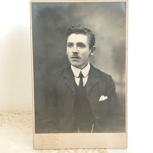 May include: A vintage black and white cabinet card photograph featuring a portrait of a person with short dark hair and a mustache, wearing a pinstriped suit jacket, a dark vest, a white collared shirt, and a tie. The text Webber The Grand Studio Lancaster is embossed at the bottom.