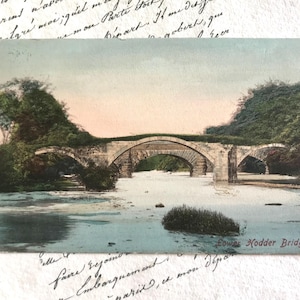 May include: Vintage postcard featuring a stone bridge with multiple arches over a river. Lush green trees line the banks, and the sky has a soft pink hue. The text "Lower Hodder Bridges" is visible.
