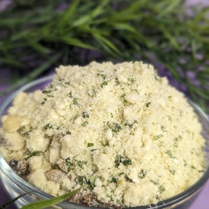 May include: A clear glass bowl filled with a mixture of dried herbs and spices. The mixture is a light beige color with green flecks throughout. The bowl is sitting on a purple surface.