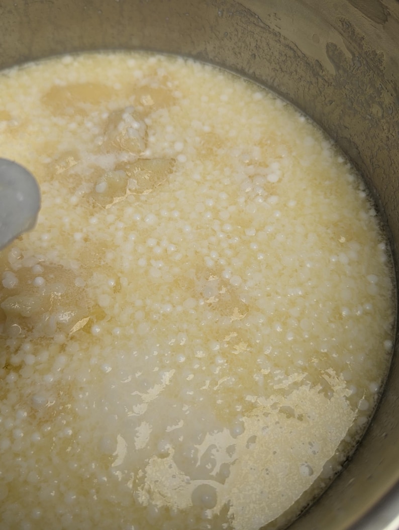 May include: Close-up of a pot filled with a creamy, off-white liquid. Small, white, spherical objects are suspended throughout the liquid, creating a textured appearance. The pot's interior is visible, with a metallic sheen.