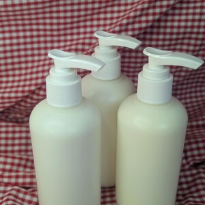 May include: Three off-white lotion or soap dispenser bottles with white pump tops. The bottles are cylindrical and sit on a red and white checkered fabric background. The dispensers are filled with a creamy liquid.