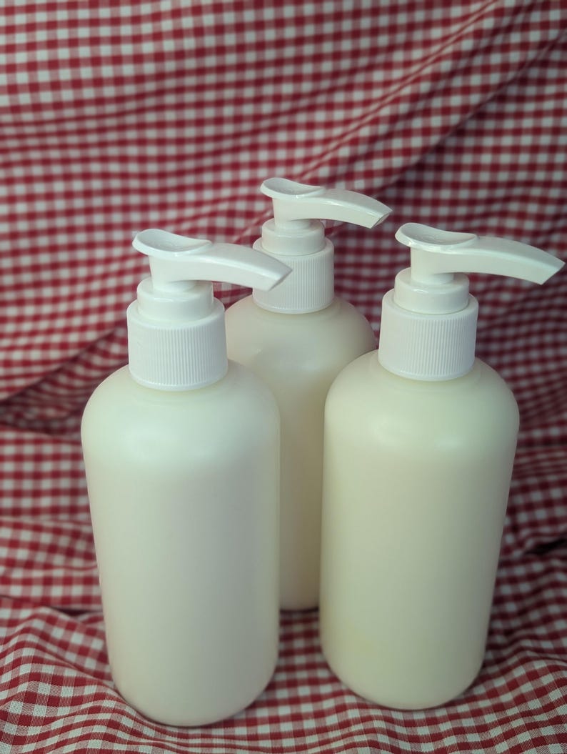 May include: Three off-white lotion or soap dispenser bottles with white pump tops. The bottles are cylindrical and sit on a red and white checkered fabric background. The dispensers are filled with a creamy liquid.
