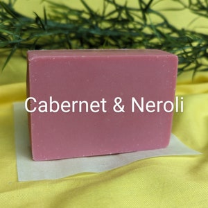 May include: A rectangular bar of soap with the text "Cabernet & Neroli" printed on the front. The soap is a light pink color and is sitting on a white piece of paper.
