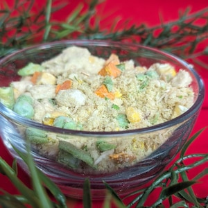 May include: A clear glass bowl filled with a mixture of dried vegetables and grains. The ingredients include green peas, orange carrots, and yellow corn, with a light brown, granular base. The bowl sits on a red surface, with green foliage in the foreground.