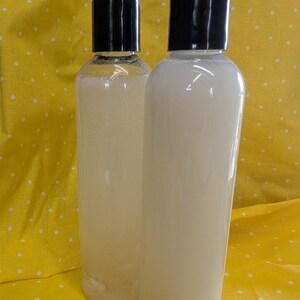 May include: Two clear plastic bottles filled with a light-colored liquid, each topped with a black cap. The bottles are cylindrical and appear to contain a cosmetic product, set against a yellow background with white polka dots.