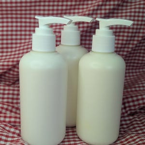 May include: Three white lotion or soap dispensers with white pump tops. The dispensers are cylindrical and filled with a creamy liquid. They are arranged on a red and white checkered fabric background.