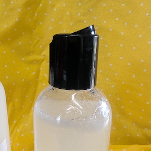 May include: A clear plastic bottle with a black flip-top cap, filled with a light-colored liquid. The bottle is partially visible against a yellow background with white polka dots. The cap is open, revealing the bottle's contents.