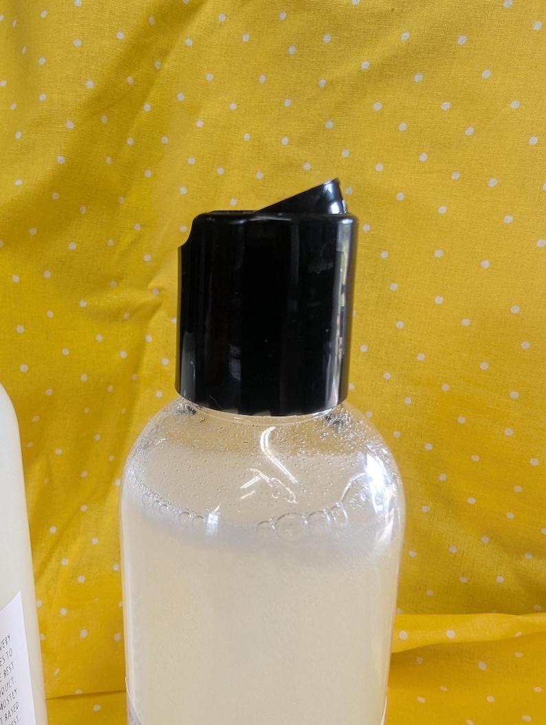 May include: A clear plastic bottle with a black flip-top cap, filled with a light-colored liquid. The bottle is partially visible against a yellow background with white polka dots. The cap is open, revealing the bottle's contents.