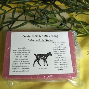May include: A bar of Goats Milk & Tallow Soap in Cabernet & Neroli. The soap is a deep red color and is wrapped in clear plastic. The label is white with black text and a black and white illustration of a goat. The label includes the ingredients and the maker's information.