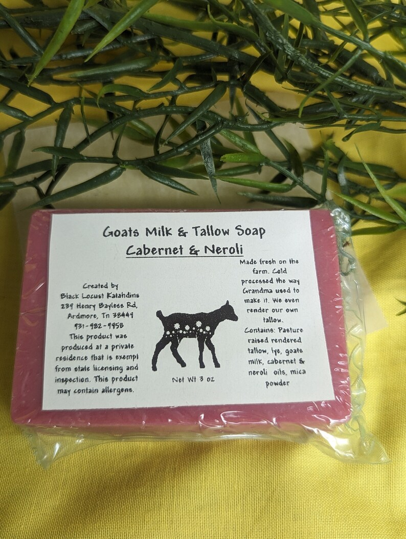 May include: A bar of Goats Milk & Tallow Soap in Cabernet & Neroli. The soap is a deep red color and is wrapped in clear plastic. The label is white with black text and a black and white illustration of a goat. The label includes the ingredients and the maker's information.