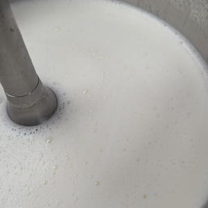 May include: Close-up of a stainless steel immersion blender in a white liquid, likely a dairy product or a sauce, inside a metal container. The liquid has small bubbles, indicating it's been recently mixed.
