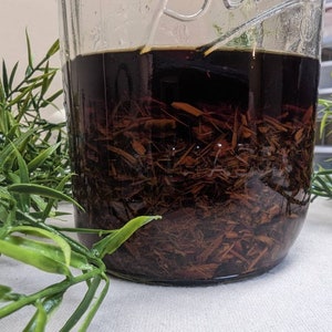 May include: A clear glass jar filled with a dark brown liquid and brown plant material. The jar is sitting on a white surface.