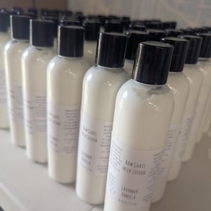 May include: A collection of white lotion bottles with black caps. Each bottle has a label that reads "Raw Goats Milk Lotion, Lavender Vanilla". The bottles are arranged on a white surface.