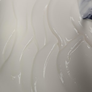 May include: Close-up of a smooth, white, creamy substance with a glossy sheen. The texture appears thick and flowing, with gentle curves and highlights. The image suggests a cosmetic product or food item.
