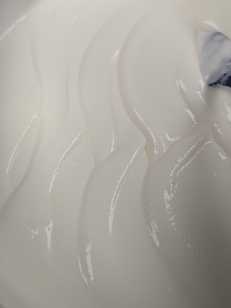May include: Close-up of a smooth, white, creamy substance with a glossy sheen. The texture appears thick and flowing, with gentle curves and highlights. The image suggests a cosmetic product or food item.