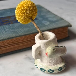 May include: A small, white ceramic vase with a snake wrapped around it, holding a yellow flower. The vase has a green and white geometric pattern at the base. A pink alarm clock and a blue book are in the background.