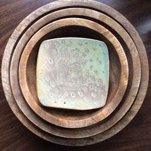 Könnte beinhalten: Ein Satz von vier Holzschalen unterschiedlicher Größe, die ineinander verschachtelt sind. In der Mitte befindet sich ein kleiner, quadratischer Keramikteller mit hellgrüner und blauer Glasur. Der Keramikteller hat ein dekoratives Design.