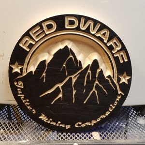 May include: A round wooden sign with a black background and gold lettering. The sign says "Red Dwarf Jupiter Mining Corporation" and features a stylized image of mountains.