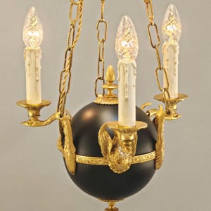Könnte beinhalten: Ein dreiarmiger Kronleuchter mit einer zentralen mattschwarzen Kugel, die mit verzierten goldfarbenen Schwanenmotiven akzentuiert ist. Die Leuchte umfasst drei kerzenförmige Glühbirnen mit weißen Tropfeffekt-Hülsen an einer goldfarbenen Kette.