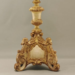 Può includere: Un candeliere alto e ornato con una finitura color oro e crema. La base è ampia e decorativa con volute, che porta a uno stelo sottile e intagliato con dettagli scanalati e una piattaforma circolare superiore per sostenere una candela.