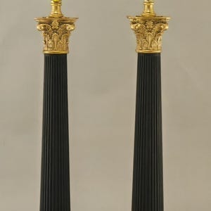 Op de afbeelding: Een paar decoratieve tafellampen in zwart en goud. Elke lamp heeft een zwarte, gegroefde kolom, een goudkleurig, sierlijk kapiteel en een gouden lamphouder. De basis is vierkant en goudkleurig, met een decoratieve rand.