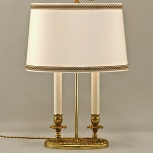 Op de afbeelding: Een tafellamp met een goudkleurige metalen voet, twee kaarsvormige fittingen en een ovale witte stoffen kap. De lamp is bekroond met een decoratieve gouden knop in de vorm van een ring met een pijl die naar rechts wijst.