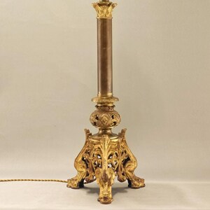 Può includere: Una lampada da tavolo dorata con un design ornamentale. La lampada ha una base rotonda con tre gambe e una colonna cilindrica alta. Il paralume è mancante.