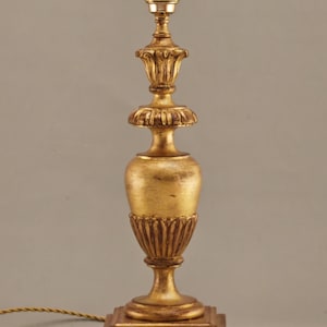 Può includere: Lampada da tavolo color oro con un design ornato. La lampada ha una base quadrata, un corpo a forma di urna e una sezione superiore dettagliata con un apparecchio di illuminazione. L'altezza della lampada è di circa 46 cm.