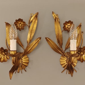 May include: A pair of gold-toned metal wall sconces featuring floral and leaf motifs. Each sconce holds a single clear, textured, flame-shaped light bulb set within a white candle-style sleeve, surrounded by sculpted metal petals and foliage.