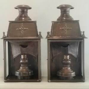 Può includere: Una coppia di lanterne da carrozza in metallo marrone scuro abbinate, caratterizzate da una cornice rettangolare, una parte superiore affusolata con un emblema a croce decorativo e un serbatoio centrale per bruciatore a olio visibile.