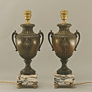 Op de afbeelding: Twee bronzen urnvormige tafellampen. Elke lamp heeft een decoratief ontwerp, gebogen handvatten en staat op een marmeren voet met goudkleurige poten. De lampen hebben goudkleurige lamphouders en zijn bedraad.