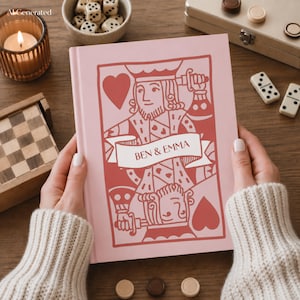 Puede incluir: Un libro de tapa dura rosa claro con una ilustración gráfica roja de un rey de naipes y una cinta en el centro con el texto BEN & EMMA. El diseño incluye símbolos de corazones y una figura de rey reflejada con una espada.