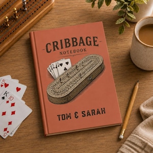 Könnte beinhalten: Ein terrakottafarbenes Notizbuch mit dem aufgedruckten Text "CRIBBAGE NOTEBOOK" und "TOM & SARAH" auf dem Einband. Der Einband zeigt eine Illustration eines Cribbage-Bretts aus Holz mit Metallstiften und Spielkarten, die zwei Fünfen zeigen.