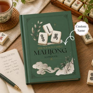 Puede incluir: Un libro de puntuación de tapa dura en color verde oscuro con el texto The Wilson Family Mahjong Scorebook en la portada. La cubierta presenta ilustraciones de tres fichas de mahjong, nubes estilizadas y tallos de bambú.