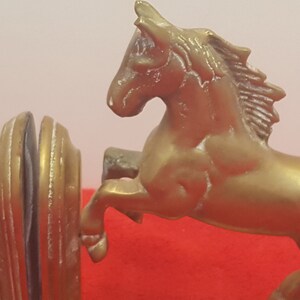 May include: A pair of gold-colored metal bookends featuring rearing horses. Each bookend displays a horse in a dynamic jumping pose with a textured mane and tail, attached to a rounded, vertical support base with a smooth, polished finish.