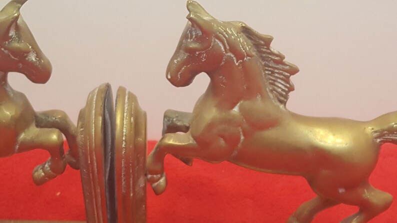 May include: A pair of gold-colored metal bookends featuring rearing horses. Each bookend displays a horse in a dynamic jumping pose with a textured mane and tail, attached to a rounded, vertical support base with a smooth, polished finish.