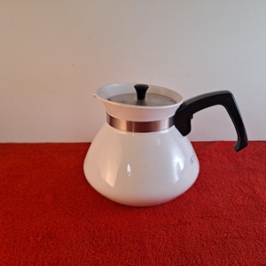 May include: White ceramic coffee pot with a black handle and a silver band around the top. The pot is sitting on a red surface.
