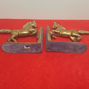May include: A pair of brass bookends featuring two horses in a rearing position. Each horse is attached to a vertical support plate and a flat, rounded base. The metal has a polished, reflective gold-toned finish with visible texture on the manes.