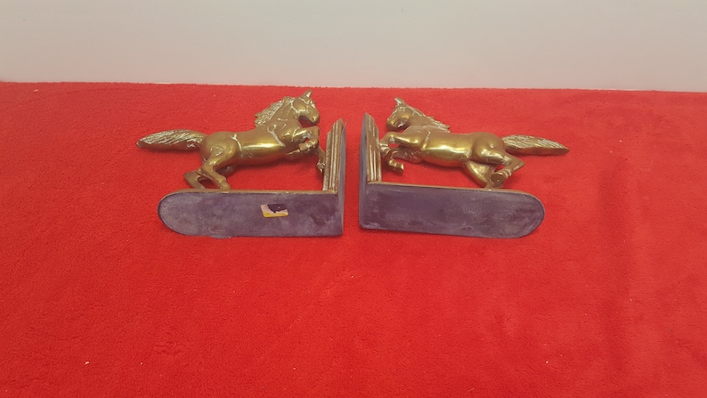 May include: A pair of brass bookends featuring two horses in a rearing position. Each horse is attached to a vertical support plate and a flat, rounded base. The metal has a polished, reflective gold-toned finish with visible texture on the manes.