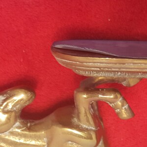 May include: A brass horse figurine supporting a horizontal shelf. The horse is depicted in a rearing position with its front legs raised. The shelf has a smooth, flat top surface and a metallic edge, showing a polished gold-toned finish throughout.
