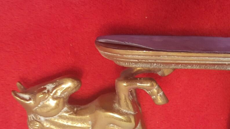 May include: A brass horse figurine supporting a horizontal shelf. The horse is depicted in a rearing position with its front legs raised. The shelf has a smooth, flat top surface and a metallic edge, showing a polished gold-toned finish throughout.