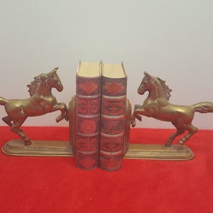 May include: A pair of brass bookends featuring rearing horses on rectangular bases, supporting two vintage-style books with dark red covers, intricate gold-toned embossed patterns, and visible page edges between the metal horse figures.