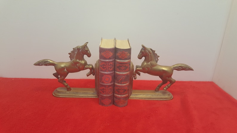 May include: A pair of brass bookends featuring rearing horses on rectangular bases, supporting two vintage-style books with dark red covers, intricate gold-toned embossed patterns, and visible page edges between the metal horse figures.