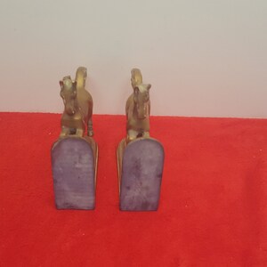 May include: A pair of gold-colored metal horse bookends. Each bookend features a standing horse figure mounted on a vertical base with a dark, flat, felt-covered bottom surface. The horses are depicted in a forward-facing stance with tails raised.
