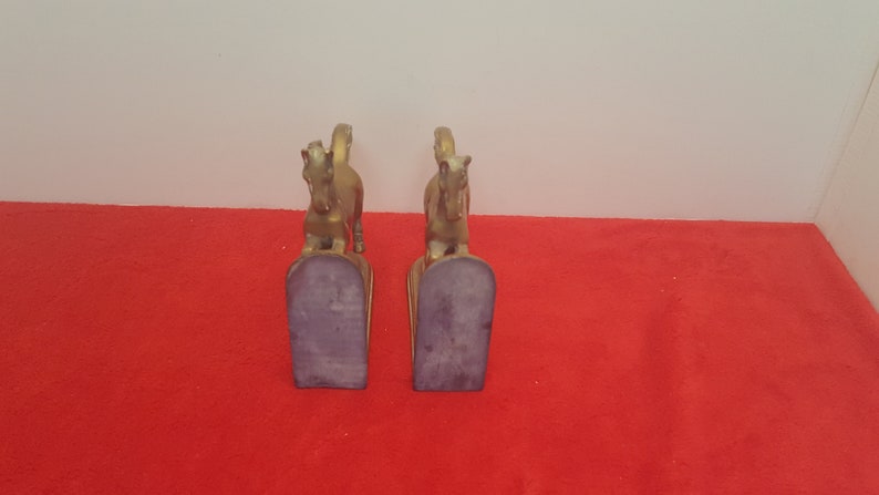 May include: A pair of gold-colored metal horse bookends. Each bookend features a standing horse figure mounted on a vertical base with a dark, flat, felt-covered bottom surface. The horses are depicted in a forward-facing stance with tails raised.