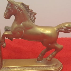 May include: A pair of gold-colored metal bookends featuring a horse in a rearing position. Each bookend has a solid base and a sculpted horse figure with a flowing mane and tail. The metal has a smooth, reflective finish throughout the design.