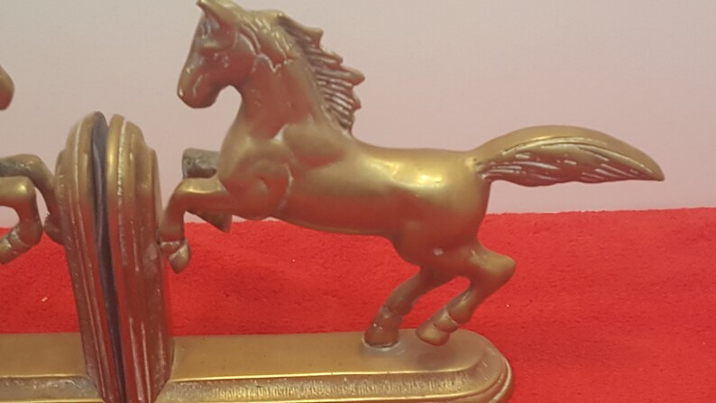 May include: A pair of gold-colored metal bookends featuring a horse in a rearing position. Each bookend has a solid base and a sculpted horse figure with a flowing mane and tail. The metal has a smooth, reflective finish throughout the design.