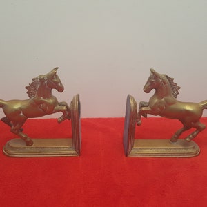 May include: A pair of brass bookends featuring two horses in a rearing position. Each horse is attached to a flat, vertical base plate with a stepped edge design. The metal has a warm, metallic gold tone with visible texture on the manes and tails.