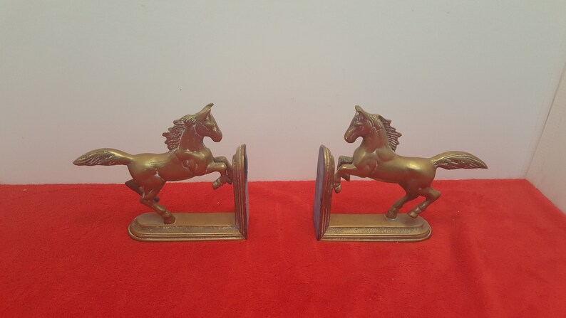 May include: A pair of brass bookends featuring two horses in a rearing position. Each horse is attached to a flat, vertical base plate with a stepped edge design. The metal has a warm, metallic gold tone with visible texture on the manes and tails.